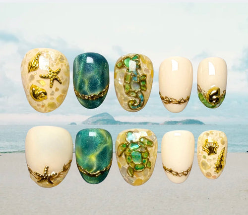 Ten press-on nails arranged in two rows against a beach backdrop. The nails feature sea-themed designs including starfish, seahorse, turtle, shell, and coral motifs in gold, green, blue, and cream tones in a shell mosaic pattern. 
