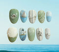 Ten decorative press-on nails arranged in two rows against a light blue sky and ocean background. The nails feature pastel green, cream, white, and blue designs with nautical motifs including a seashell, sailboat, lighthouse, waves, and silver shell and pearl embellishments.