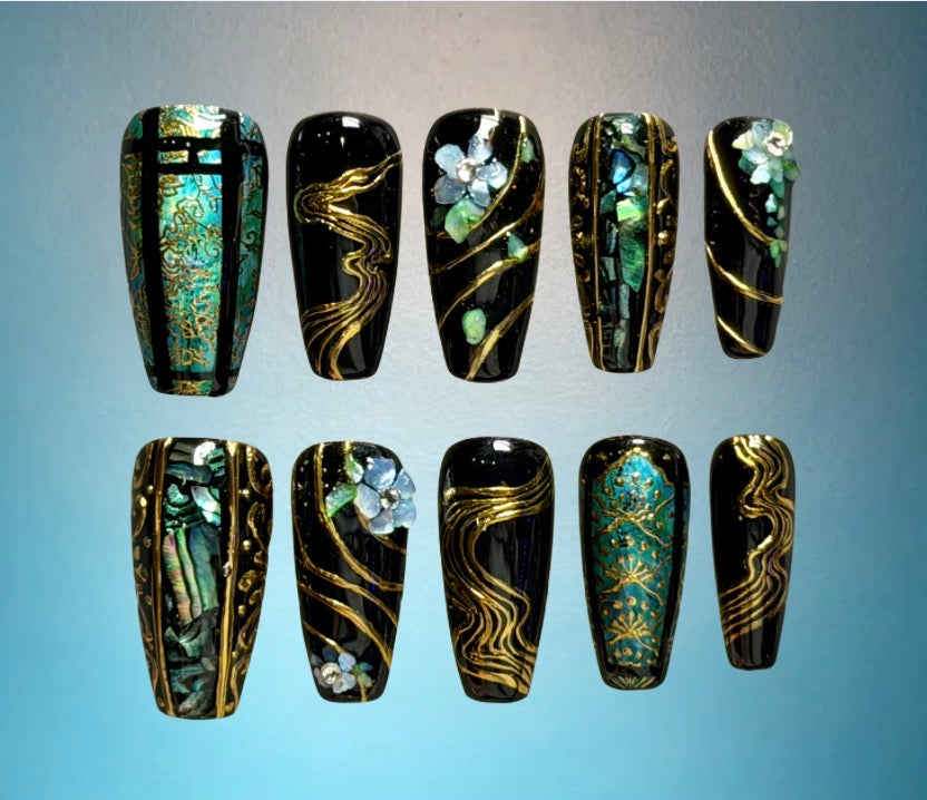 Ten coffin-shaped press-on nails arranged in two rows on a light blue gradient background. The nails feature glossy black and turquoise bases with intricate designs including gold wavy lines and floral accents, decorations using abalone shell