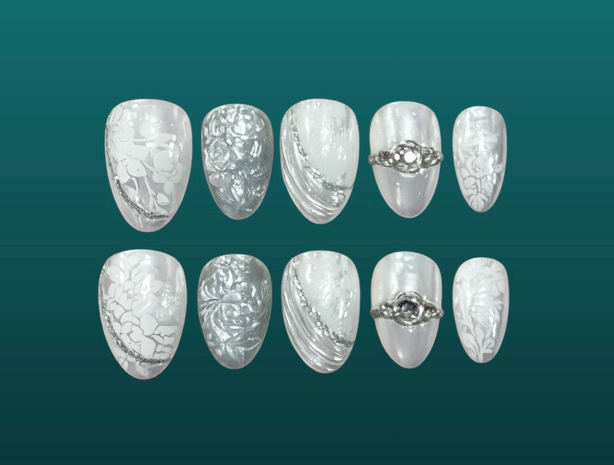 Ten press-on nails in a bridal design with white lace florals, silvery satin drapery, and engagement ring flourishes on a shining white silver base. 