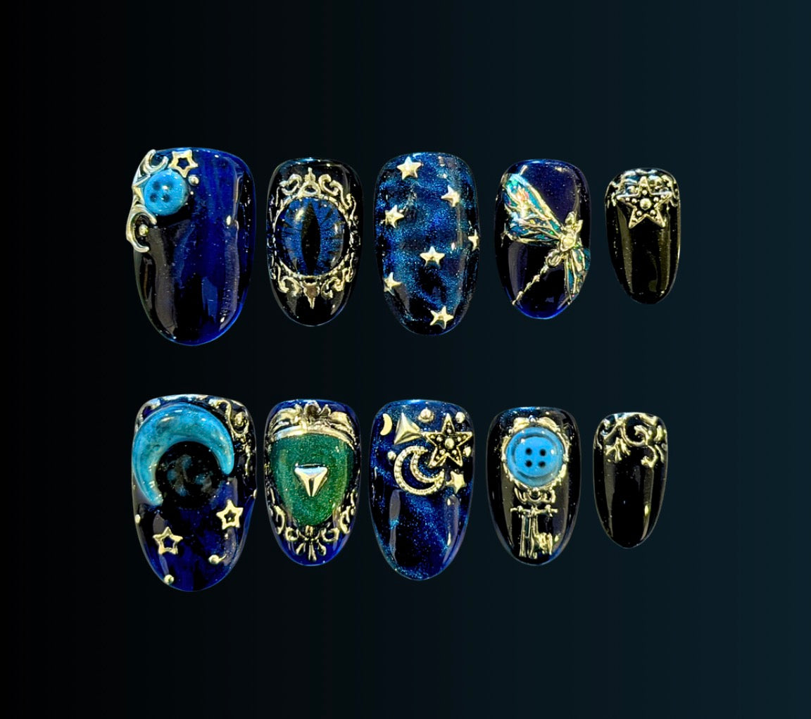Coraline-inspired press-on nail set in midnight blue with antique silver filigree, celestial stars, button and key details, cat-eye accents, and hidden glow-in-the-dark elements.